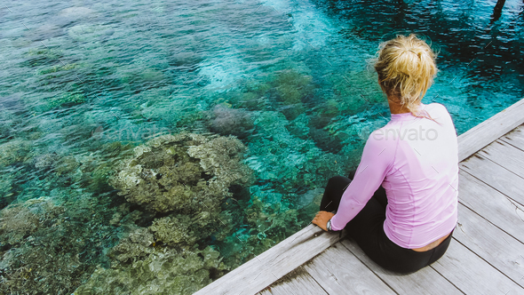 Girl sitting on a wooden jetty near Mansuar island in Raja Ampat. Beautiful colorful corals visible - Stock Photo - Images