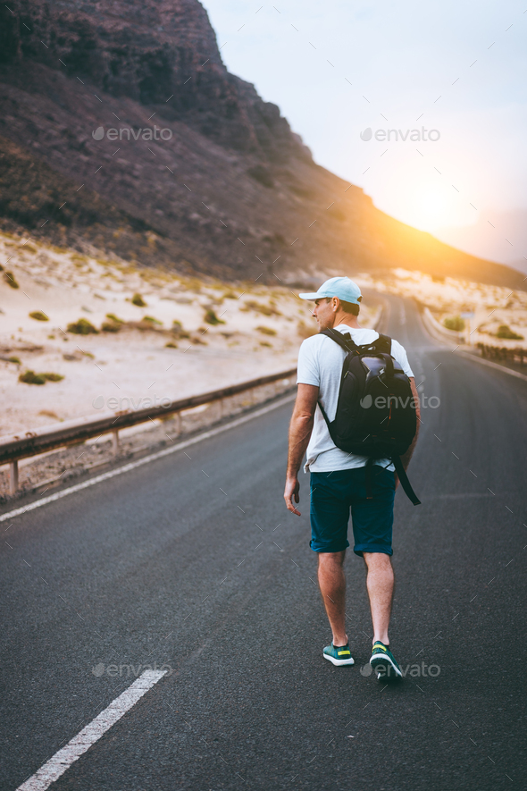 Traveler with backpack walks in the center of an epic winding road. Huge volcanic mountains in the - Stock Photo - Images