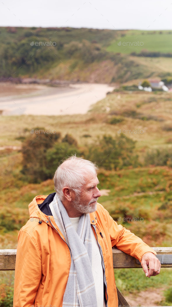 Active Senior Man Walking Along Coastal Path In Fall Or Winter By Gate With Beach And Cliffs Behind