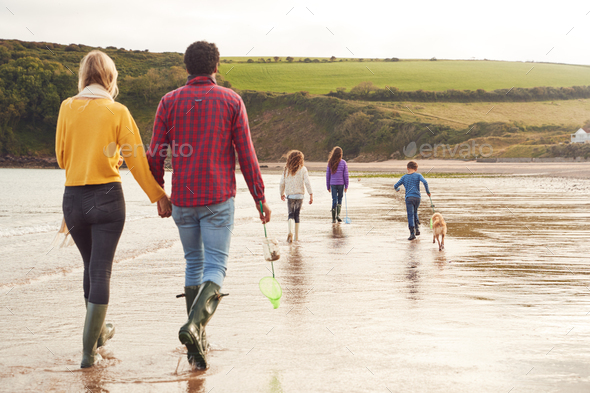 Rear View Of Multi-Cultural Family With Pet Dog Walking Along Beach Shoreline On Winter Vacation