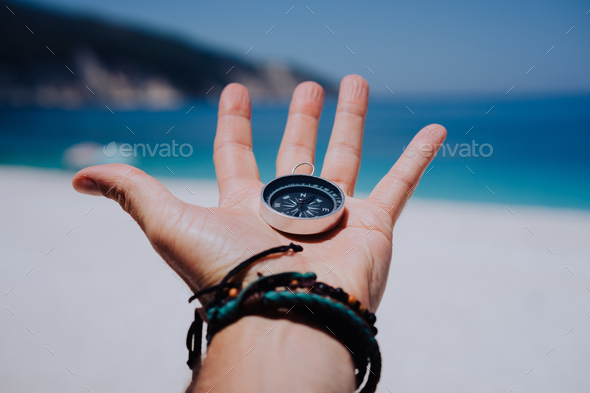Outstretched hand holding black metal compass against white sandy beach and blue sea. Find your way