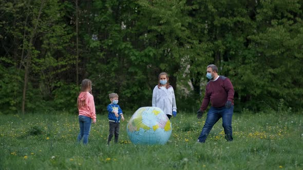 A Father with Children in Medical Masks Plays with a Large Inflatable Ball in the Park in the Spring