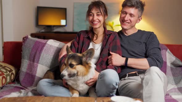 Happy Couple with Cute Pet Welsh Corgi Dog Sitting on Couch at Home at Evening alt