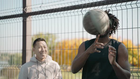 Black Man Spins a Basketball on His Finger with His Friend Smiling on a Court alt