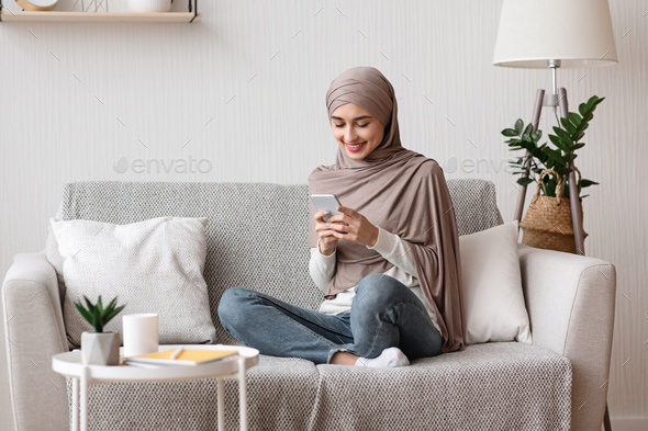 Happy arabic woman in hijab using smartphone on sofa at home - Stock Photo - Images