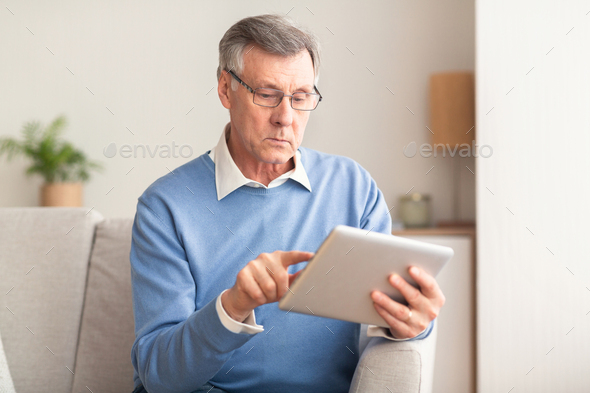 Elderly Man Using Tablet Browsing Internet Sitting On Couch Indoor