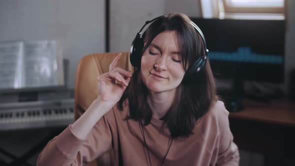 A beautiful girl with headphones listens to music in a chair at the computer and sings, smiles.
