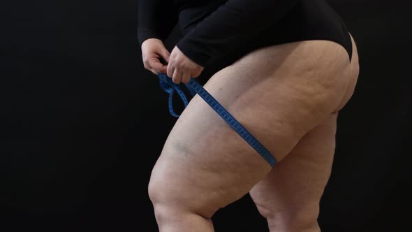 a Fat Woman with Cellulite Measures Herself with a Measuring Tape on a Black Background