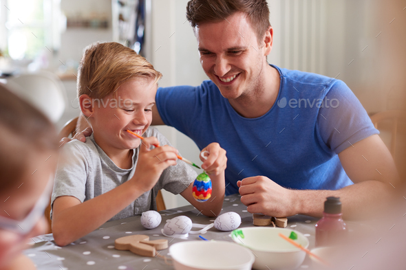 Father With Children Sitting At Table Decorating Eggs For Easter At Home