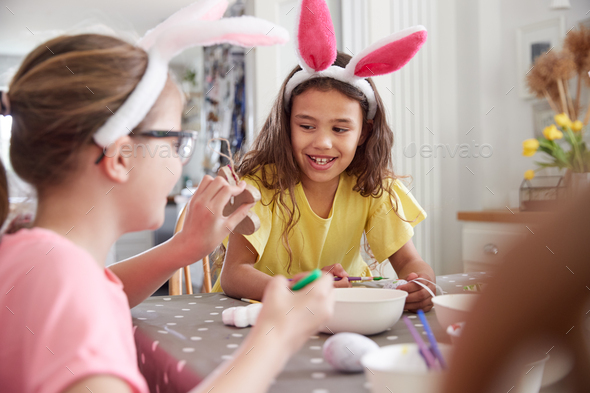 Three Girls Wearing Bunny Ears Sitting At Table Decorating Eggs For Easter At Home