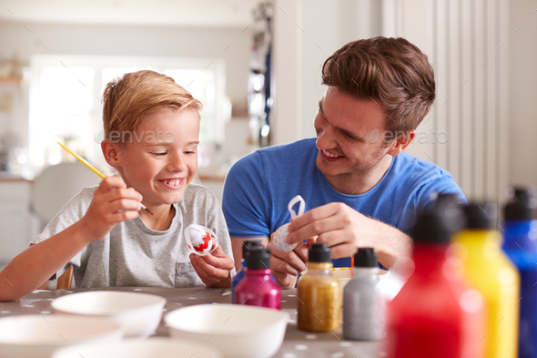 Father With Son Sitting At Table Decorating Eggs For Easter At Home