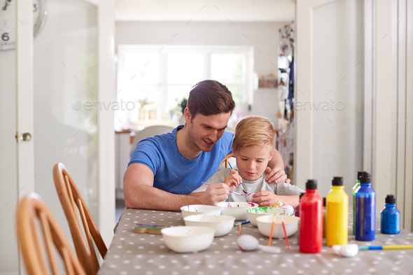 Father With Son Sitting At Table Decorating Eggs For Easter At Home