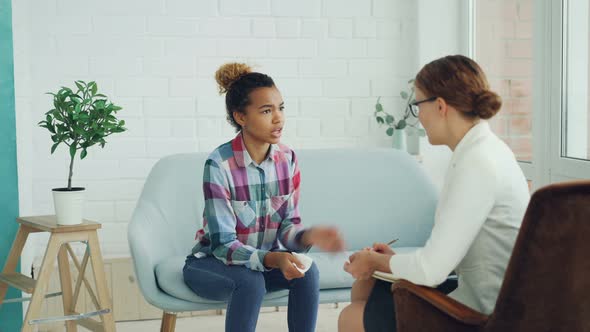 Upset Young Lady in Casual Clothing Is Having Therapy Session with Psychologist in Light Studio alt