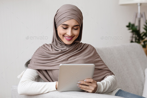 Smiling muslim woman in hijab using digital tablet, relaxing at home