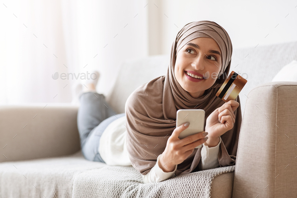 Pensive girl in hijab with smartphone and credit card lying on couch - Stock Photo - Images