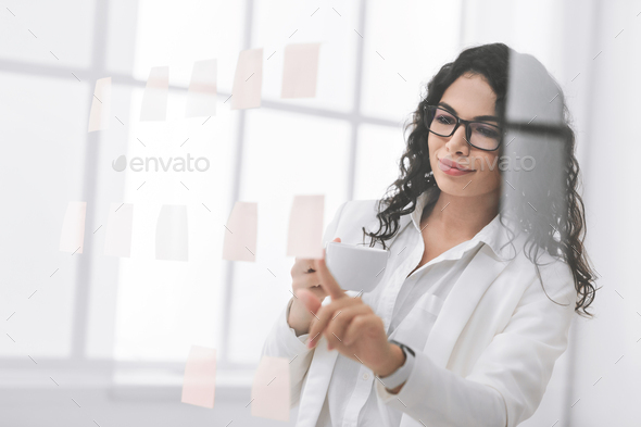 Hispanic businesswoman looking at sticky notes on glass wall