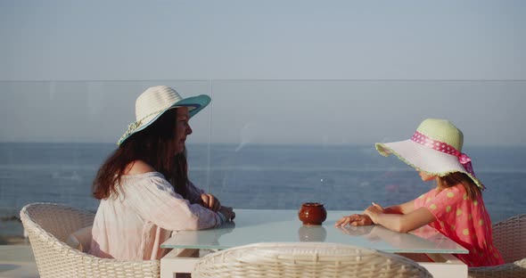 Mom and Daughter Teenager in Summer Hat Talking in Outdoor Cafe on Sea Landscape