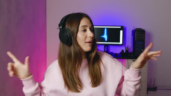Young Woman in Pink Sweatshirt in Headphones Dancing and Singing in Modern Room with Neon Background alt