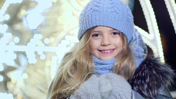 Portrait of Happy Cute Little Girl Looking at Camera on Blurred Christmas Lights Background alt
