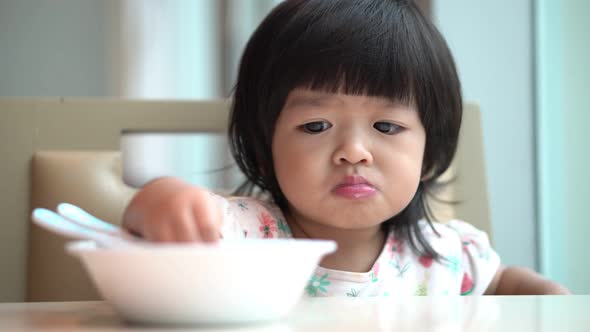 Happy Asian toddler using a spoon to scoop cereal to eat breakfast on the table in kitchen