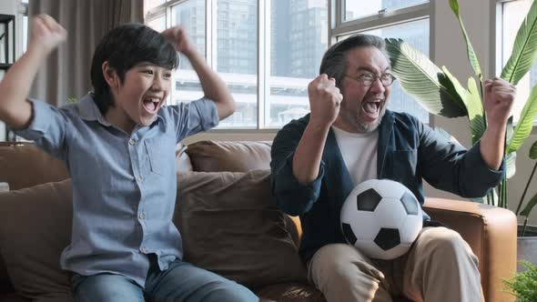 Asian father and son watching football sports games on TV Shoot the ball into the goal.