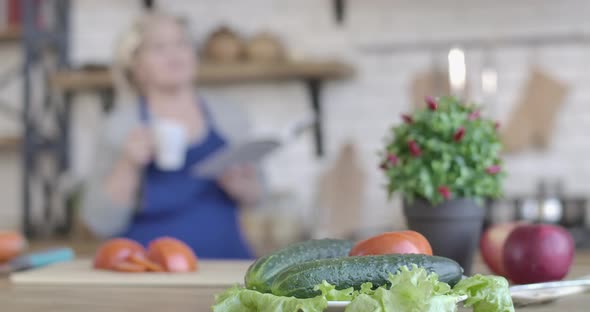 Close-up of Fresh Vegetables Lying on Table in Kitchen As Blurred Woman Drinking Coffee and Reading alt