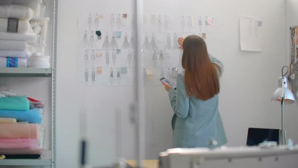 Female Fashion Designer Looking at Drawings and Sketches That are Pinned to the Wall Behind Her Desk alt