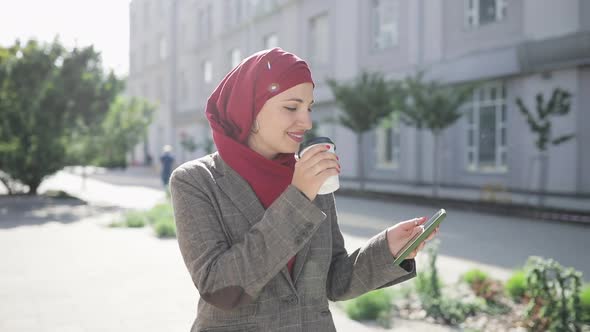 Young Muslim Woman Wearing Hijab Headscarf Walking in the City Center, Using Smartphone and Drinking