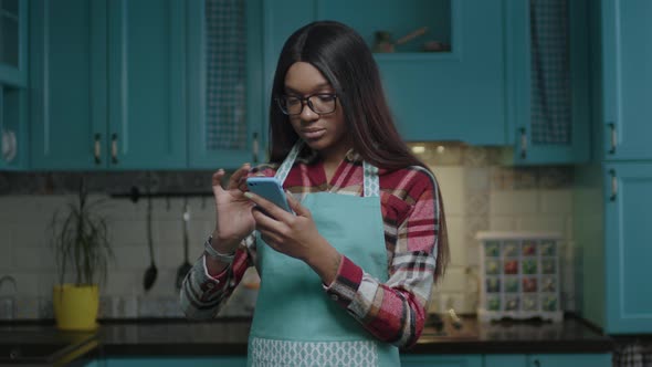 20s African American Woman in Apron Searching Online Recipe Standing on Blue Kitchen and Then alt