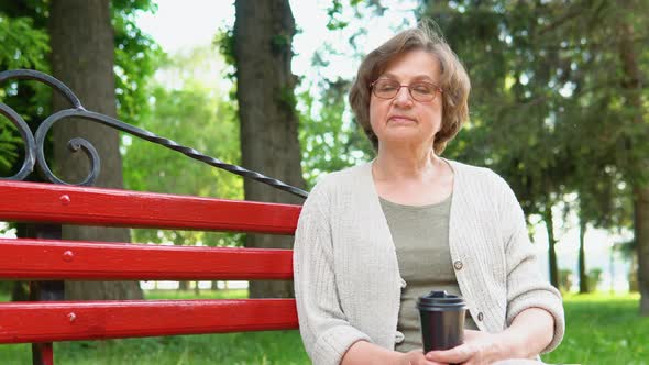 Portrait of an Older Elderly Woman with Coffee Sitting in a Park on a Bench alt