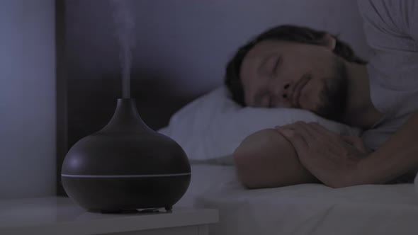 At Night Man Slips in Bed with an Aromatherapy Diffuser Working on a Nightstand alt