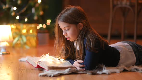 little girl lies on the floor against the background of a festive Christmas tree and reads a book.