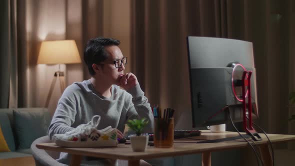 Asian Male Footwear Designer Thinking While Working On A Desktop Computer At Home alt