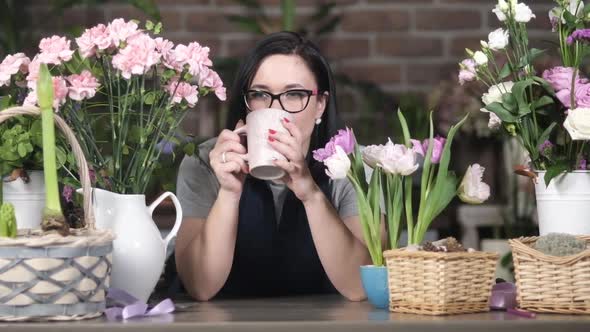 Young girl florist drinks coffee among blooming roses, hyacinths and carnations. alt