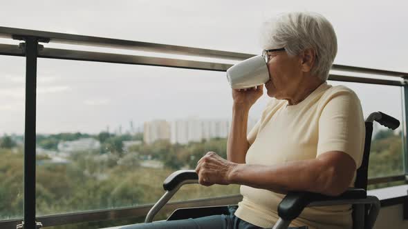 Senior Gray Haired Woman with Eyeglasses Drinking Cup of Coffee in the Wheelchair in Front alt