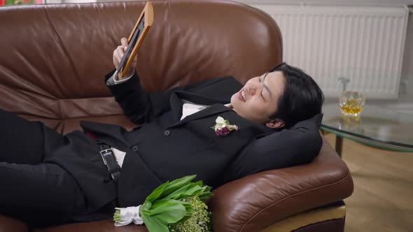 Portrait of Happy Young Asian Man in Wedding Suit Lying on Couch Admiring Picture in Hand alt
