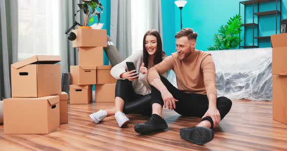 Young Couple are Using Phone Amid Boxes in a New House alt