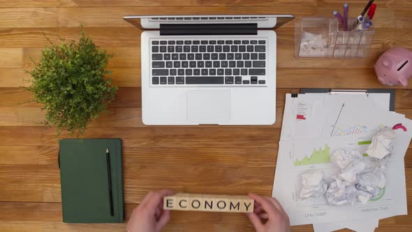 Word Economy is Made Up of Wooden Cubes on an Office Table Next to Work Documents and a Laptop alt
