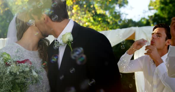 Bride and groom kissing while guest blowing bubbles alt