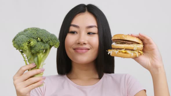 Asian Woman Holding Burger And Broccoli Choosing Food White Background alt