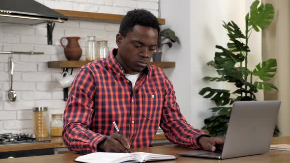 African American Man Freelancer Works for Laptop in Kitchen Writes in Notebook alt