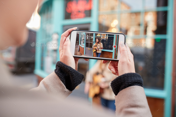 Person Filming Female Musician Busking Playing Acoustic Guitar And Singing To Crowd On Mobile Phone
