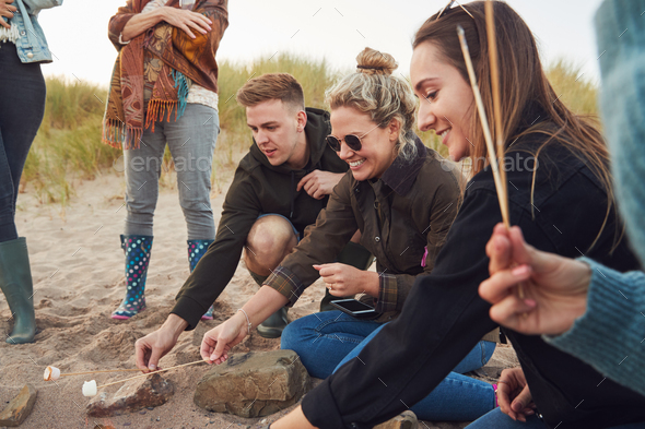 Group Of Young Friends Toasting Marshmallows Around Fire On Beach Vacation
