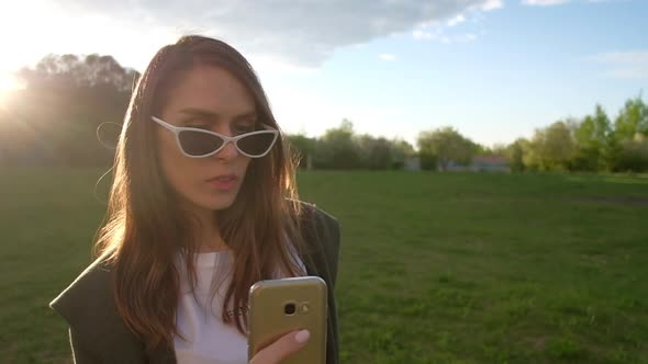 Beautiful Young Woman Making Selfie with Golden Smartphone in a Park alt