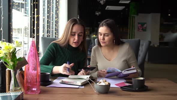 Two Young Businesswomen Are Sitting in a Cafe and Talking To a Partner on a Speaker