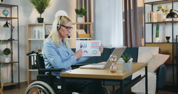 Woman in Headphones Sitting in Wheeelchair and Holding Online Meeting with Coworkers alt