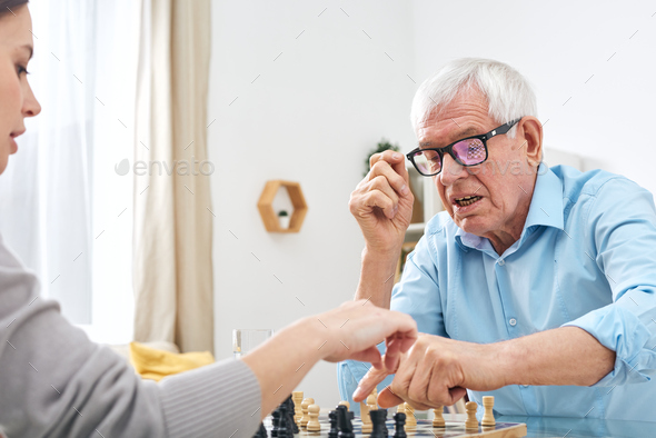 Senior disable man in eyeglasses and shirt playing chess with social worker