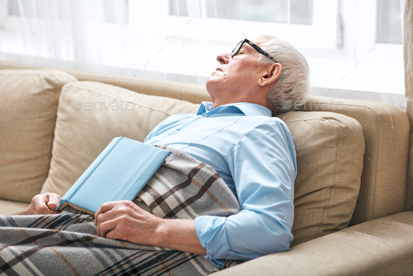 Tired senior man covered with plaid napping on couch with open book