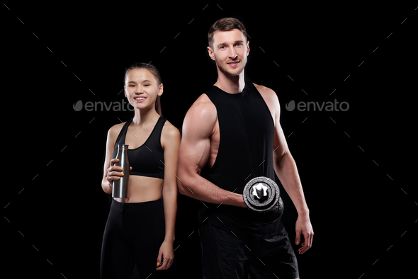 Happy young sportswoman with bottle of water and muscular man with barbell