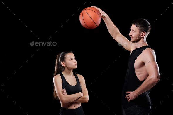 Young muscular basketball trainer holding ball over fit sportswoman